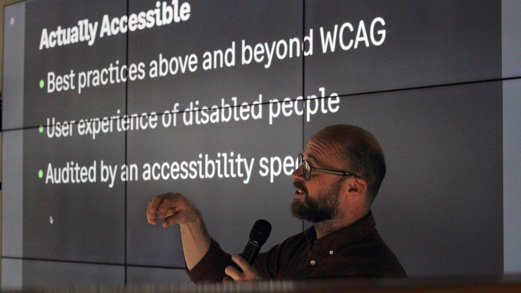 A man with a microphone talking in an friendly, animated manner; the slide on the big screen behind him is about accessibility being more than WCAG compliance.