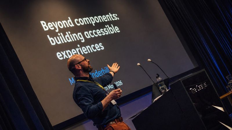 A profile shot of Martin Underhill on stage, gesturing towards the slide projected behind him with his open left hand. The slide reads ‘Beyond components: building accessible experiences'.