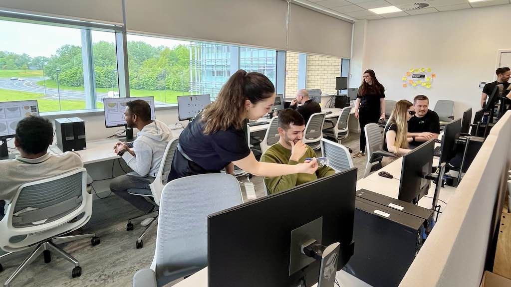 A room full of desktop computers with five people trying out simulators as three accessibility champions in black t-shirts guide them.