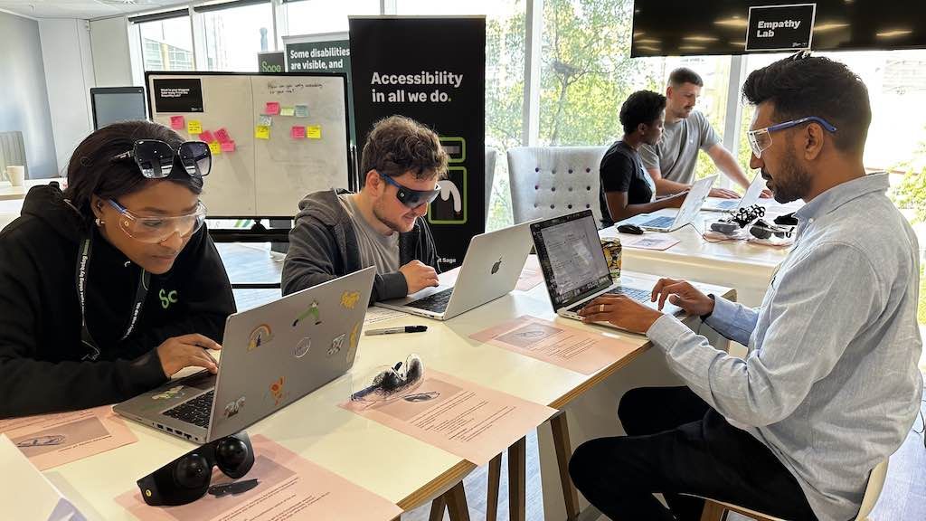 Three people in the foreground wearing visual impairment goggles trying out tasks on their laptops using the worksheets next to them. In the background, two more people are talking and looking at a laptop.