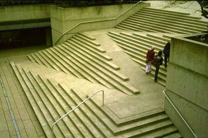 A set of stairs that has a ramp zigzagging back and forth cutting down the steps. The steps are frequently interrupted by the ramp and there are no handrails except for at the side of the stairs.