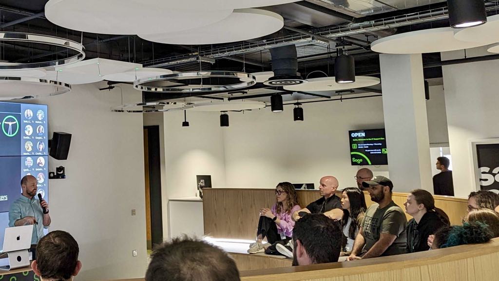 A man presenting in front of a large screen, showing a Teams call with lots of people dialled in; there is also a group of people gathered round in the room itself to watch.